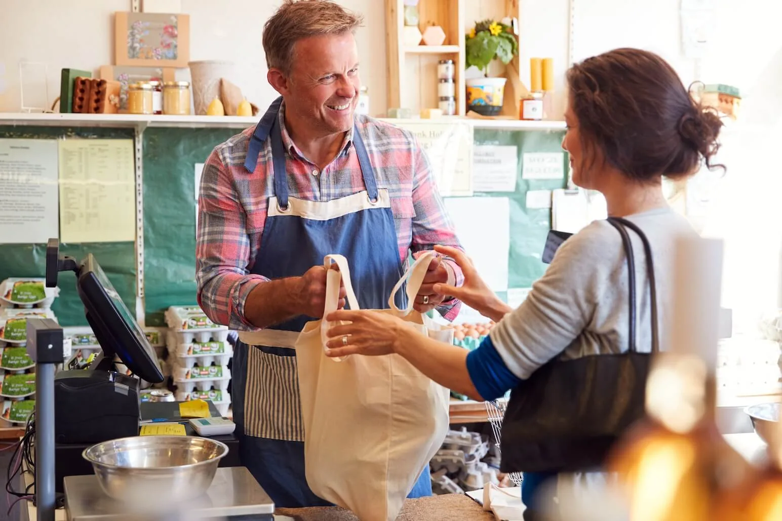 Local shop owner in an apron handing a canvas bag to a customer across the counter